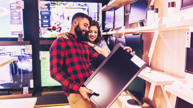 Casal feliz analisando uma televisão em suas mãos, dentro de uma loja de eletrodomésticos.