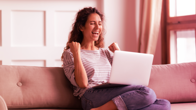 Mulher feliz olhando para o notebook com os punhos cerrados comemorando.