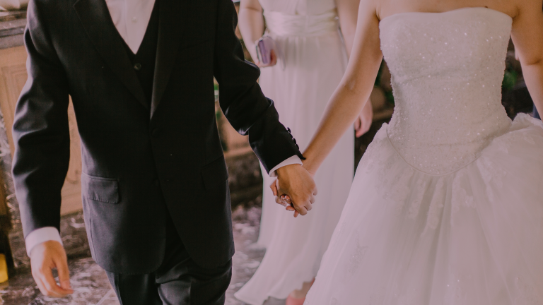 Casal de noivos de pele branca de mãos dadas no casamento. O noivo está com um terno preto, colete preto e camisa branca, a noiva está com um vestido branco