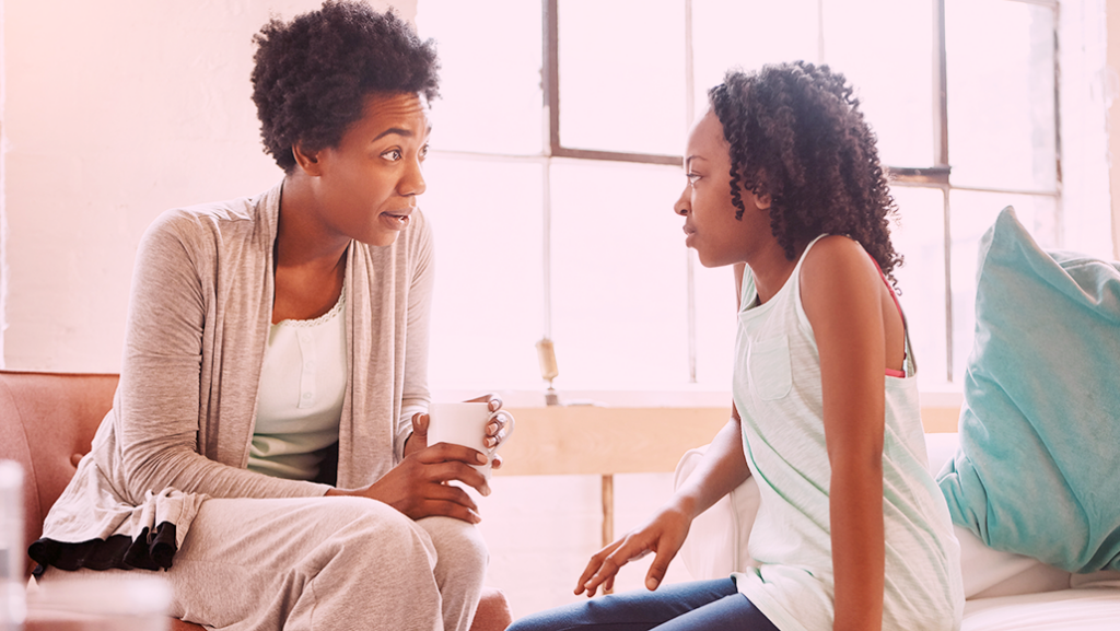 Uma mulher e uma criança negras sentadas de frente uma pra outra conversando. A mulher explica algo pra criança e está segurando uma caneca.