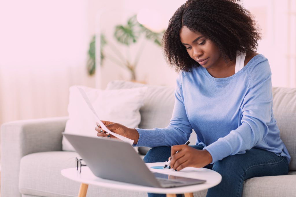 mulher sentada em um sofá. Ha uma mesa de centro a sua frente na qual ela apoia um notebook e faz anotações em uma folha com uma caneta enquanto segura uma folha