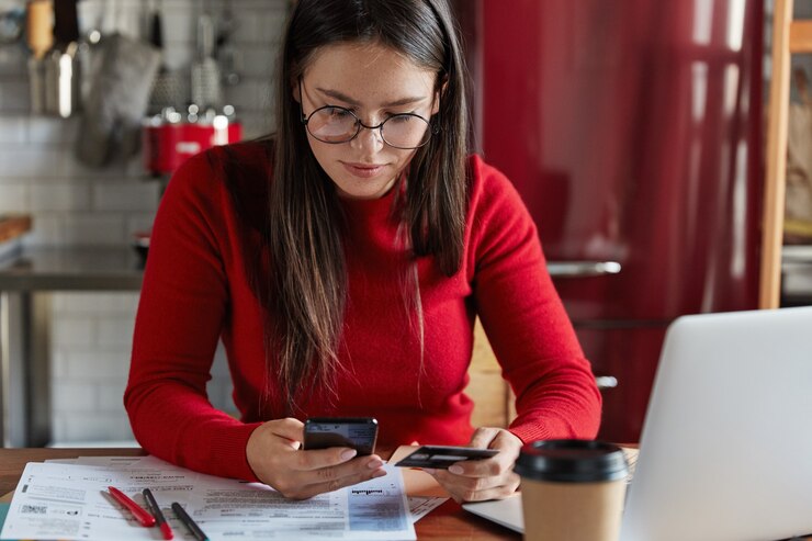 Mulher de vermelho em uma mesa olhando para seu celular e segurando um cartão de crédito