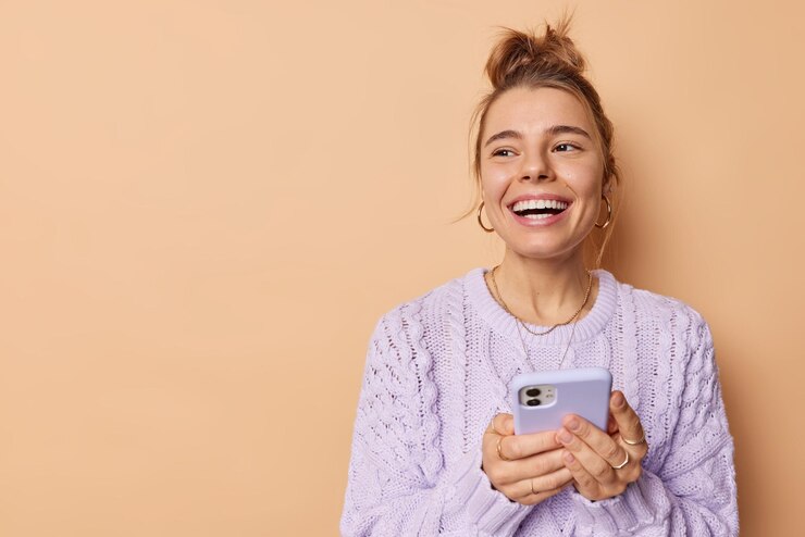 Sorridente mulher europeia positiva com cabelo penteado usa celular gosta de tecnologias modernas mantém telefone celular usa suéter casual isolado pesquisando sobre open finance